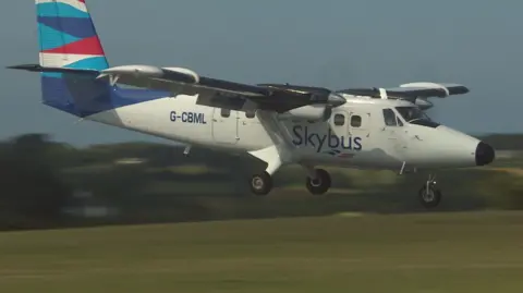 A Skybus aircraft with twin propellers prepares to land at an airport. The aircraft is mostly white in colour with a blue, red and white tailfin. The company logo is on the side of the plane. It's call sign G-CBML is written in blue block capitals on the side of the aircraft.