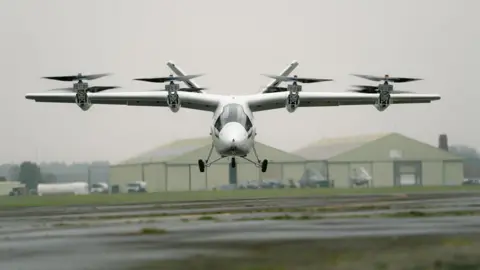 The aircraft seen from the front, with its two wings above the fuselage and eight rotors mounted front and back. Below it is a runway and on the distance there are two pale green aviation sheds. 