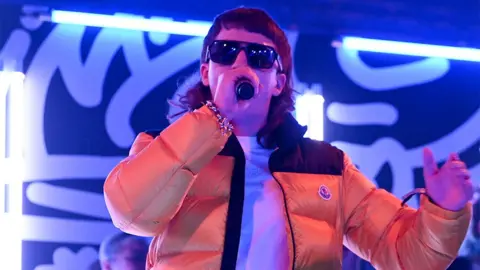 Getty Images Clive 'Sam' Robinson of the Bad Boy Chiller crew on stage, wearing an orange puffer jacket and black sunglasses, singing into a black microphone. There are neon blue lights in the background.