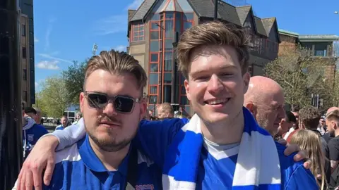 Christoffer Vårhus Two football fans stand together to pose for a photo. They wear matching blue football shirts. They both have short brown hair and one is wearing glasses.