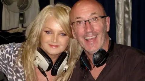 Mal Pope A woman with long blonde hair sat down next to a bald man wearing glasses. They both have headphones around their necks and smiling at the camera. 