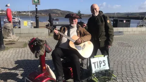 Derek Adams Derek Adams with dark green waterproof coat standing next to a man with a dark-coloured tricorn hat sitting on a paved area playing a guitar, with a sign saying "Tips" on the ground next to him. The sea is visible behind and there is a red lifebelt on a stand to the left.