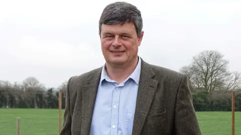 NFU Nigel Barnes stands outdoors in a large open grassy field. He is wearing a light blue button-down shirt and smart woollen jacket.
