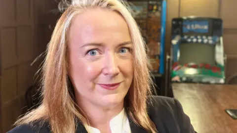 Portrait photo of Professor Lorraine Hope who has long straight brown hair with blonde highlights and blue eyes. She is smiling to camera in a dark panelled room.