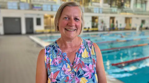 BBC/Edward Sault Annette wearing a sleeveless blue floral top stands in front of an indoor swimming pool. She is centered in the frame and facing the camera. The background is softly blurred and shows multiple swimming lanes, poolside decking, and a bright, modern aquatic facility with large windows and upper-level walkways.