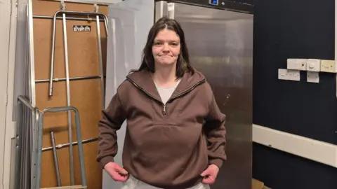 Sionne stands smiling to the camera, wearing a brown jumper inside the cafe. A large fridge is stood behind.