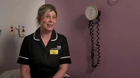 A nurse in uniform sits on a bed in the corner of a purple coloured room. Equipment can be seen in the room behind her.
