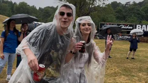 Connor Bennett/BBC A man and a woman in transparent rain shawls. The woman is giving a peace/V-sign. There is a bar behind them in the park.