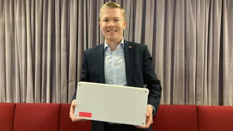 BBC Bradley Thomas MP, a man with short blond hair is wearing a dark jacket over a pale blue shirt. He is standing in front of a line of red chairs with grey curtains behind them and holding a white box with "streetwave" embossed on the side.