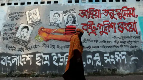Students-painted graffiti on a grey wall near Dhaka University area, including illustrations of four female rape victims. The Bengali writing on the walls demands justice for all rapes, including those involving children. A woman in a black robe and orange hijab is seen walking past it.