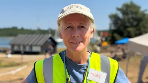 Image shows Mary Montagu-Scott smiling at the camera. She has a 'Shipyard Trust" cap on. She is wearing a yellow high-vis and blue top. Behind her, is a black boat house.