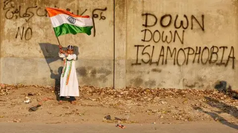 Getty Images A young boy stand holding the national flag next to a wall that says 'Down down Islamophobia'