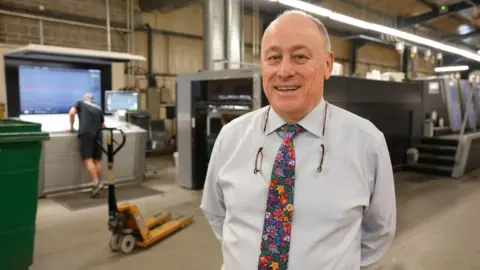 Andrew in a white shirt and floral tie smiles as he stands in an industrial warehouse, with machinery and a worker blurred in the background