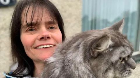 A woman with black hair smiling at camera holding a large grey cat. She is wearing a blue coat with a grey jumper underneath. The picture background is of her stone dashed house and has a window on her left.