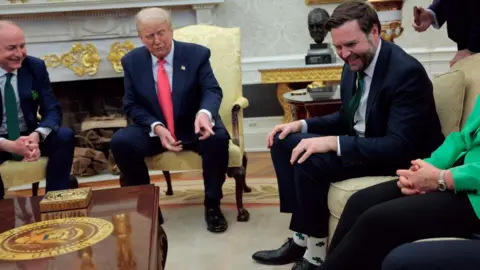 Getty Images Donald Trump señala con los calcetines del vicepresidente de EE. UU. JD Vance mientras es anfitrión de Micheál Martin en la Casa Blanca. Vance se está riendo y Martin también se ríe. Él está a la izquierda de Trump, y ambos están sentados en una silla amarilla de respaldo alto. Los tres hombres llevan trajes.