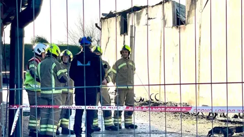 Steve Huntley/BBC Firefighters in uniform and helmets standing together, in discussion, behind a fire cordon and metal fence, beside a fire-damage farm building