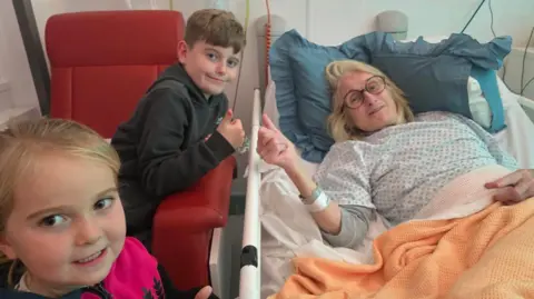 Helen in a hospital bed with her two grandchildren standing next to her. She wears a hospital gown and has a orange blanket over her. A young boy sits on a red chair next to her with his thumb up, and a young girl with blonde hair smiles at the camera. 