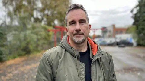 Dr Craig Johnston is wearing a green puffer jacket. He has orange lining and is wearing a black t-shirt. Dr Johnston has white and black hair and a short beard. Behind him is a Bristol street with lots of autumn leaves on the ground. 