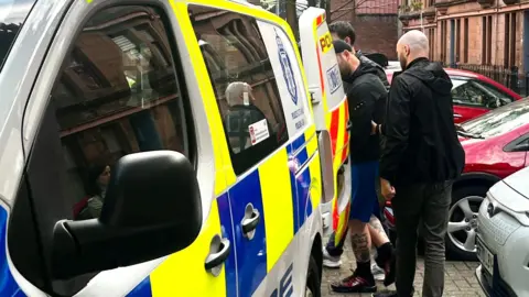 A man is escorted into the back of a blue, white and yellow Police Scotland van by two plain-clothes police officers on a residential street. Cars are parked nearby.