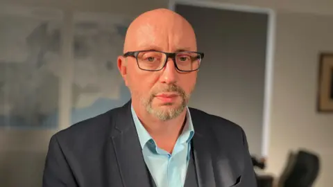 A bald man wearing glasses. He is wearing a blue shirt and looking directly at the camera. 