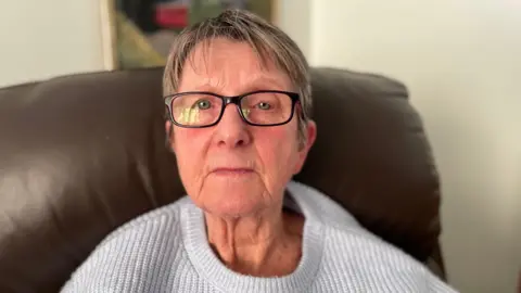Woman in her 70s with glasses and a light-coloured jumper sitting on a brown leather sofa