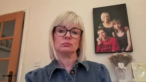 Sharron Huddleston, with blonde hair and glasses, looks at the camera with a forlorn expression. There is a photo of her with her two children on the wall behind her.