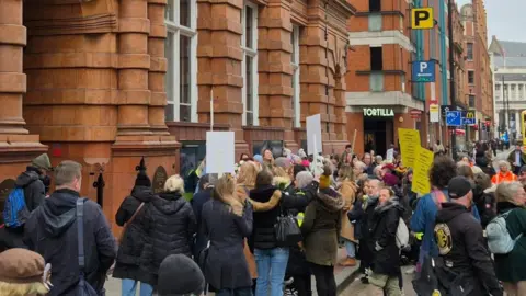 Para pengunjuk rasa Johnlawrence berkumpul di jalan dengan plakat dan tanda -tanda di luar pintu masuk bata merah ke kantor Greater Manchester Gabungan Oxford Street Otoritas.