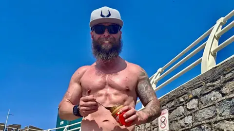 Russ Penwell wearing a hat and sunglasses and eating peanut butter on a beach.