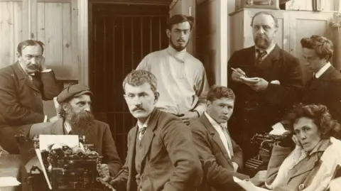 Alex McKinstry Sepia photograph of seven men and one woman sitting next to typewriters.