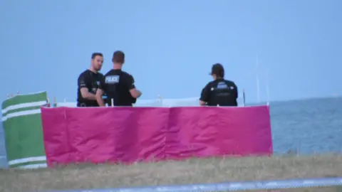 Lewis Adams/BBC Three police officers standing behind a waist-height bright pink screen on a grass with the sea behind them. 