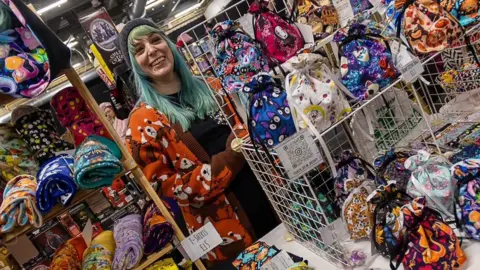 Handout Becky Forster is standing behind her That's Sew Becky stall which offers clothing and accessories. T-shirts and bags are laid out, all in bright colours. Ms Forster has dyed light green hair and is wearing a black woolly hat and smiling. The picture has been taken at an angle.
