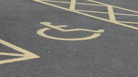 A piece of tarmac with a wheelchair symbol in yellow on the ground between two sets of yellow warning signs 