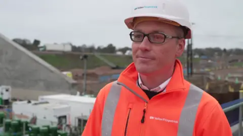Adrian Mawdsley looking at the camera has a white hard hat on, an orange hi-vi jacket and black framed glasses.