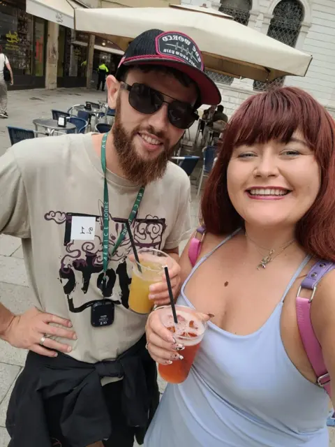 Chelsea Rees Chelsea Rees holding a drink. The mole is visible on her chest jus above her top. She is stood next to a man who has a beard and a beige t shirt on.
