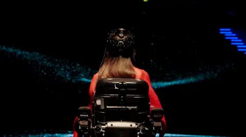 Dentsu A view taken from behind Breanna shows the back of her headset and wheelchair. Blue dots and lines from the computer visualisation appear around her.