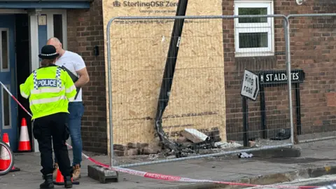 A policewoman consults with a man at the scene of the crash. A small amount of broken bricks and rubble are on the pavement. A large board covers the main damage. A road sign is knocked into a lopsided position.