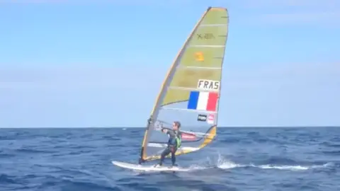 Martin Gavériaux A person is windsurfing across choppy blue sea water, riding a board with a sail that has a French flag on it.