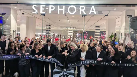 Allison Ferns A group of people in black shirts cutting the black ribbon in front of the glass-fronted shop, which says "Sephora" in white letters.