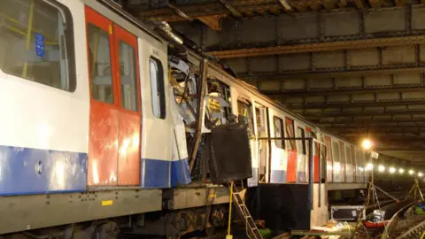 Metropolitan Police A blue and white London underground tube train with red doors is pictured underground. Part of the carriages are damaged in the middle of the train with a door and metal hanging out. A light and a ladder can be seen pointing into this section of the train with other equipment on the train tracks.