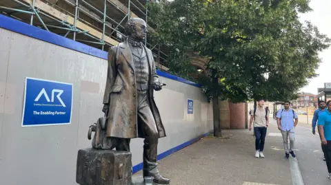A bronze statue of Thomas Cook, outside a building covered with scaffolding. Passers by look on, and a tree stands in the background of the image.