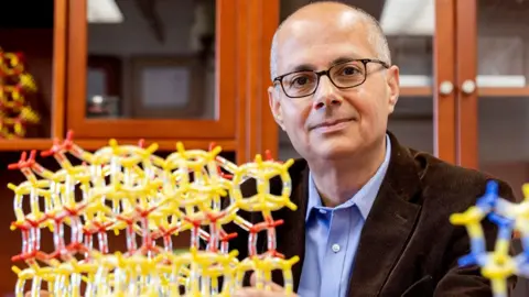 Reuters UC Berkeley chemistry professor Omar Yaghi, wearing a blue shirt, brown jacket and glasses, poses in his lab at the University of California in Berkeley US. Next to him is a model showing metal-organic frameworks made of yellow and red tubes and nodes.