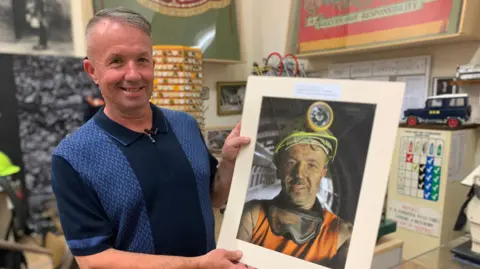 A former miner dressed in a blue shirt holds a selfie photograph. It shows him underground at Thoresby Colliery. His face is blackened with dirt. He is wearing a miner's helmet with lamp and goggles around his neck.