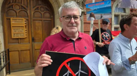 Didier Lucotte has short grey hair, is wearing glasses and a red polo shirt. He's holding a placard staring in front of large wooden doors to Tynwald behind him, and protestors to the right.