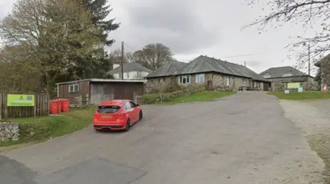 Google Street View image of YHA Dartmoor youth hostel. A red Ford Focus is driving into the driveway area of the site. Several stone buildings and a wooden shack are at the site.