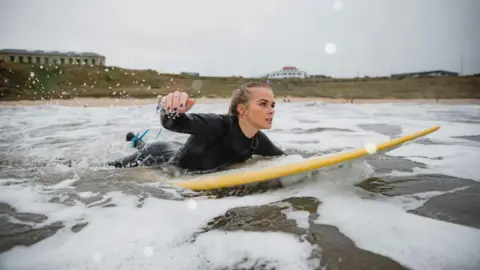 Getty Images A young woman with long blonde hair lies on a yellow surfboard and paddles out on the sea. Behind her is the beach and a cliff atop of which sits several small buildings. The picture is taken at Tynemouth.