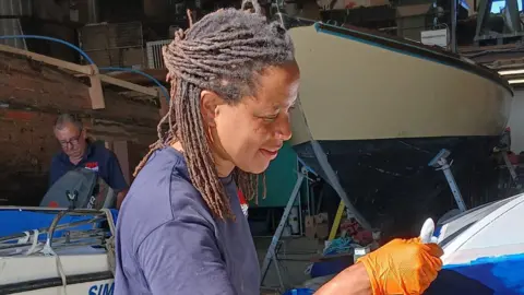 Contributed A woman paints the hull of a boat. She is smiling as she looks to the point where she is painting. She has dark braids and wears a blue T-shirt. She is also wearing orange plastic gloves. Boats are behind her as well as a man working on another vessel.