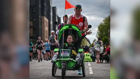 Derek Goodfellow A man in a red vest, hat and sunglasses, pushing his daughter - a young girl with brown hair and a black top - in a specialist green run chair. They are both smiling, the girl is waving.
There are numerous runners following behind them.
