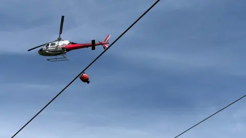 A red and white helicopter in the sky with a red water bucket hanging underneath it.