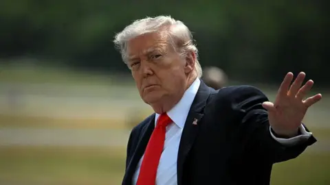 Trump with his hand up in the air waving. He wears a white shirt, red tie and black suit jacket. The background is blurry but he is outside and appears to be in front of grass 