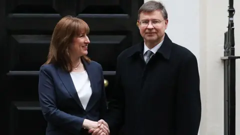 Alishia Abodunde/Getty Images Rachel Reeves, wearing a blue jacket, and Valdis Dombrovskis, wearing a black coat, shake hands while standing in front of a black door.